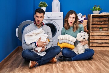 Young hispanic couple doing laundry sitting on the floor in shock face, looking skeptical and sarcastic, surprised with open mouth
