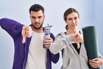 Young hispanic couple wearing sportswear with angry face, negative sign showing dislike with thumbs down, rejection concept