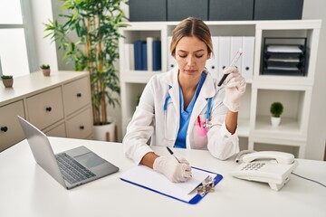 Young hispanic woman wearing doctor uniform analysing blood test tube at clinic