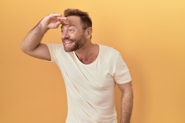 Middle age man with beard standing over yellow background very happy and smiling looking far away with hand over head. searching concept.