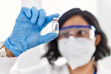 Young latin woman wearing scientist uniform and medical mask holding dose vaccine at laboratory