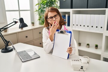 Young doctor woman wearing stethoscope holding clipboard covering mouth with hand, shocked and...