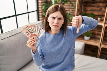 Young brunette woman holding colombian pesos with angry face, negative sign showing dislike with...