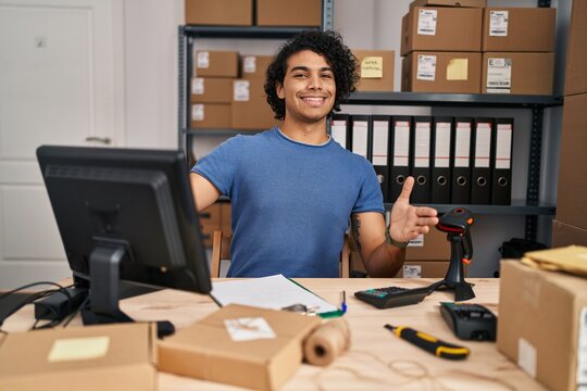Hispanic man with curly hair working at small business ecommerce looking at the camera smiling with open arms for hug. cheerful expression embracing happiness.