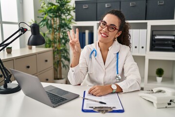Young hispanic woman wearing doctor uniform and stethoscope showing and pointing up with fingers...
