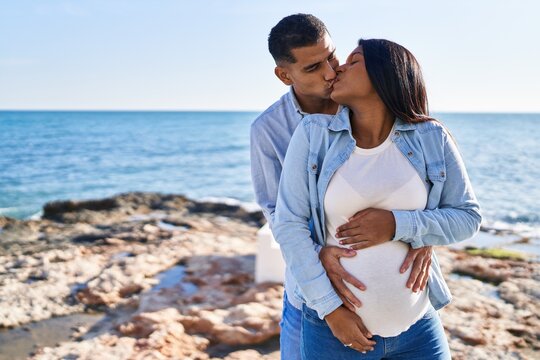 Young latin couple expecting baby hugging each other and kissing at seaside
