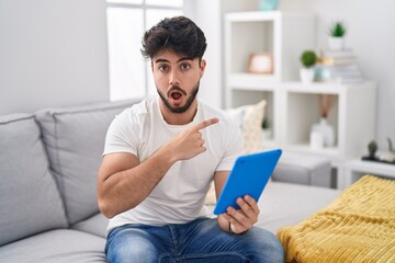 Hispanic man with beard using touchpad sitting on the sofa surprised pointing with finger to the side, open mouth amazed expression.