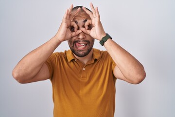 Fototapeta premium Hispanic man with beard standing over white background doing ok gesture like binoculars sticking tongue out, eyes looking through fingers. crazy expression.