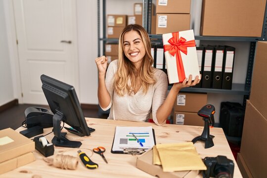 Young Blonde Woman Working At Small Business Ecommerce Holding Gift Screaming Proud, Celebrating Victory And Success Very Excited With Raised Arm