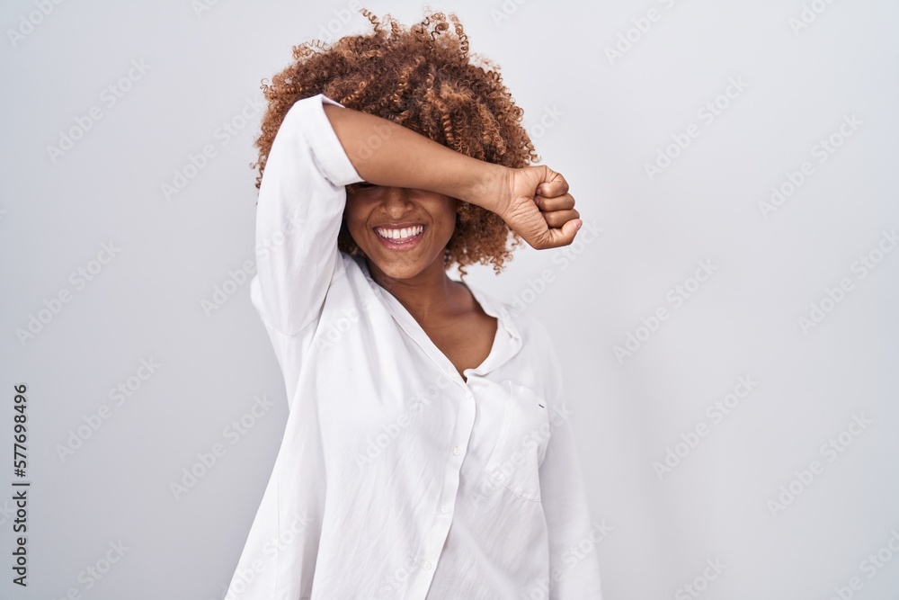 Wall mural Young hispanic woman with curly hair standing over white background covering eyes with arm smiling cheerful and funny. blind concept.