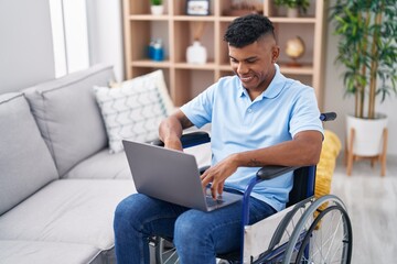 Young latin man using laptop sitting on wheelchair at home