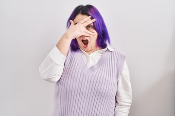 Plus size woman wit purple hair standing over white background peeking in shock covering face and eyes with hand, looking through fingers with embarrassed expression.