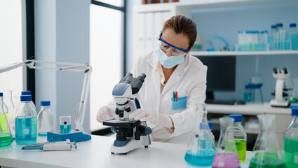 Middle age hispanic woman wearing scientist uniform and medical mask using microscope at laboratory