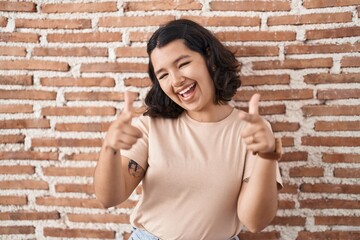 Young hispanic woman standing over bricks wall pointing fingers to camera with happy and funny face. good energy and vibes.