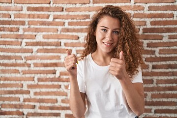 Young caucasian woman standing over bricks wall background success sign doing positive gesture with hand, thumbs up smiling and happy. cheerful expression and winner gesture.