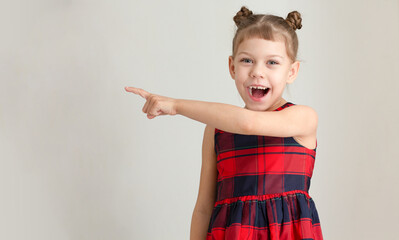 Laughing with wide open mouth child showing on copy space by index finger on one hand, caucasian kid little girl of 6 7 years in red plaid dress