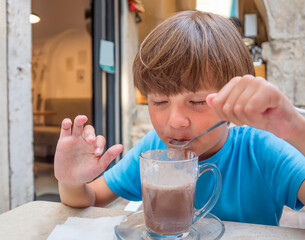 A ten-year-old Caucasian boy is sitting in a street cafe and drinking cocoa with a teaspoon.