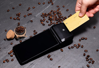 Buying coffee with gold credit card at Smart POS (point of sale) contactless. Coffee beans all over near the smart device. Black background. Smart terminal making transaction. - obrazy, fototapety, plakaty