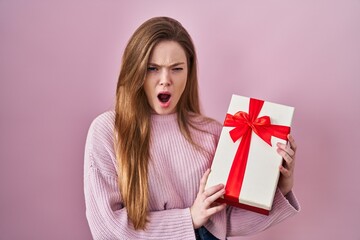 Young caucasian woman holding gift angry and mad screaming frustrated and furious, shouting with...