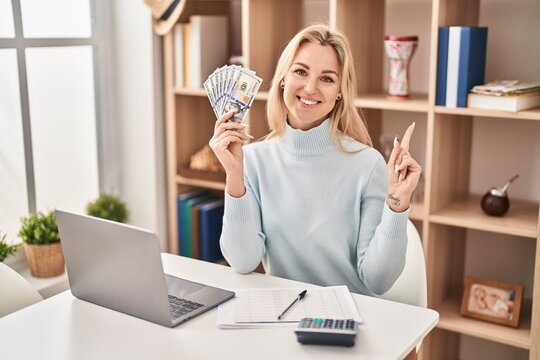 Young caucasian woman using laptop holding dollars banknotes smiling happy pointing with hand and finger to the side