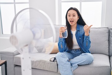 Young asian woman drinking glass of water enjoying air from fan pointing thumb up to the side smiling happy with open mouth