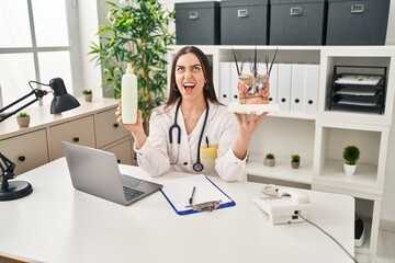 Hispanic doctor woman holding model of human anatomical skin and hair angry and mad screaming frustrated and furious, shouting with anger looking up.