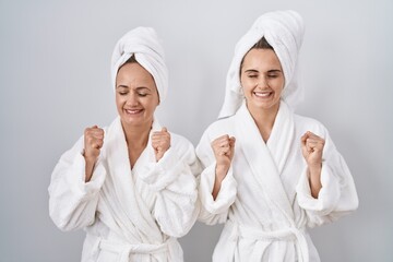 Middle age woman and daughter wearing white bathrobe and towel excited for success with arms raised...