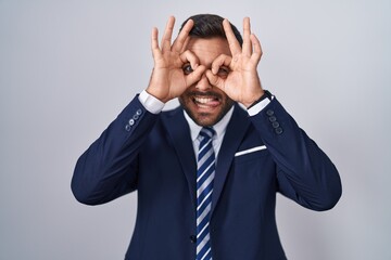 Handsome hispanic man wearing suit and tie doing ok gesture like binoculars sticking tongue out, eyes looking through fingers. crazy expression.