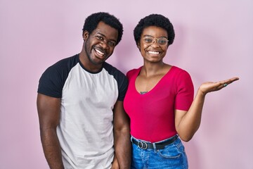 Young african american couple standing over pink background smiling cheerful presenting and pointing with palm of hand looking at the camera.