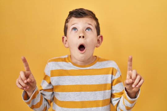 Young Caucasian Kid Standing Over Yellow Background Amazed And Surprised Looking Up And Pointing With Fingers And Raised Arms.