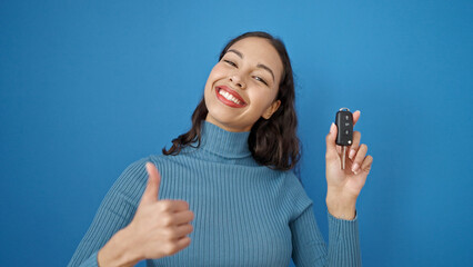 Young beautiful hispanic woman doing thumbs up holding key of new car over isolated blue background