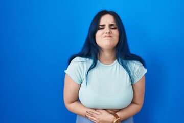Obraz premium Young modern girl with blue hair standing over blue background with hand on stomach because indigestion, painful illness feeling unwell. ache concept.