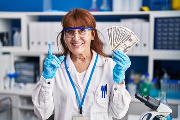 Middle age hispanic woman working at scientist laboratory holding dollars surprised with an idea or question pointing finger with happy face, number one