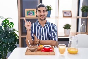 Fototapeta na wymiar Hispanic man with long hair sitting on the table having breakfast smiling happy pointing with hand and finger to the side