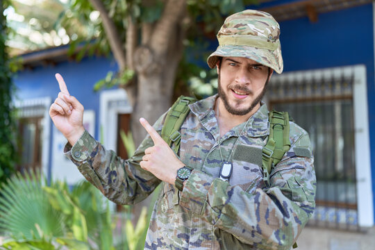Young hispanic man wearing camouflage army uniform outdoors smiling and looking at the camera pointing with two hands and fingers to the side.