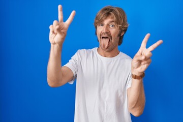 Middle age man standing over blue background smiling with tongue out showing fingers of both hands doing victory sign. number two.