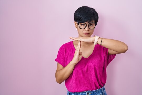 Young asian woman with short hair standing over pink background doing time out gesture with hands, frustrated and serious face