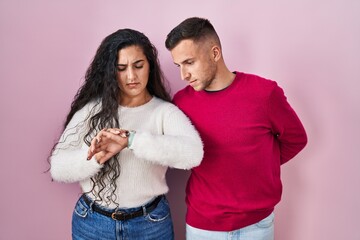 Young hispanic couple standing over pink background checking the time on wrist watch, relaxed and confident