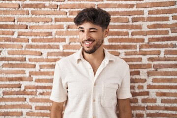 Obraz premium Arab man with beard standing over bricks wall background winking looking at the camera with sexy expression, cheerful and happy face.