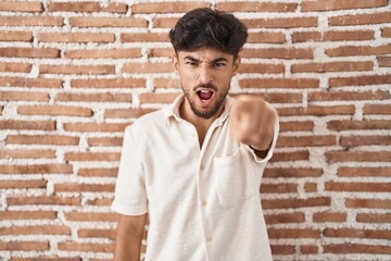 Arab man with beard standing over bricks wall background pointing displeased and frustrated to the...