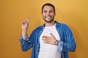 Hispanic man standing over yellow background smiling swearing with hand on chest and fingers up,...