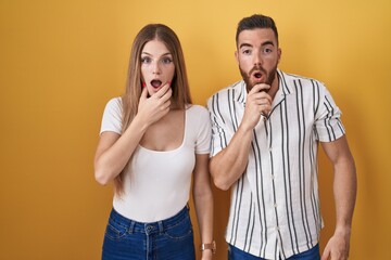 Young couple standing over yellow background looking fascinated with disbelief, surprise and amazed expression with hands on chin