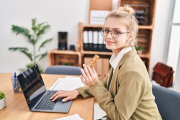 Young blonde woman business worker using laptop eating cereal bar at office