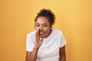 Young hispanic woman with curly hair standing over yellow background hand on mouth telling secret rumor, whispering malicious talk conversation
