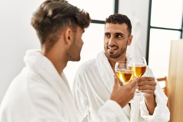 Two hispanic men couple toasting with champagne sitting on massage table at beauty center