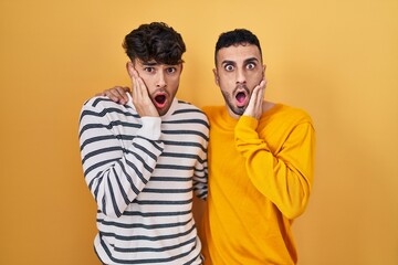 Obraz premium Young hispanic gay couple standing over yellow background afraid and shocked, surprise and amazed expression with hands on face