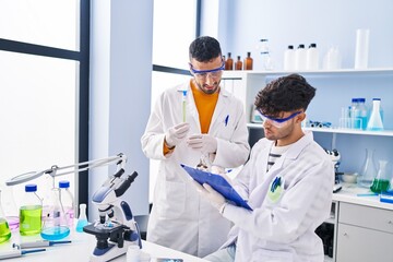 Two man scientists holding test tubes write on clipboard working at laboratory