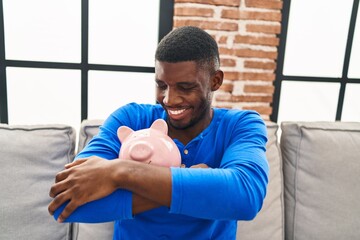 Young african american man hugging piggy bank sitting on sofa at home