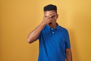 Young hispanic man standing over yellow background peeking in shock covering face and eyes with hand, looking through fingers with embarrassed expression.