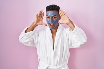 Young hispanic man wearing beauty face mask and bath robe smiling cheerful playing peek a boo with hands showing face. surprised and exited